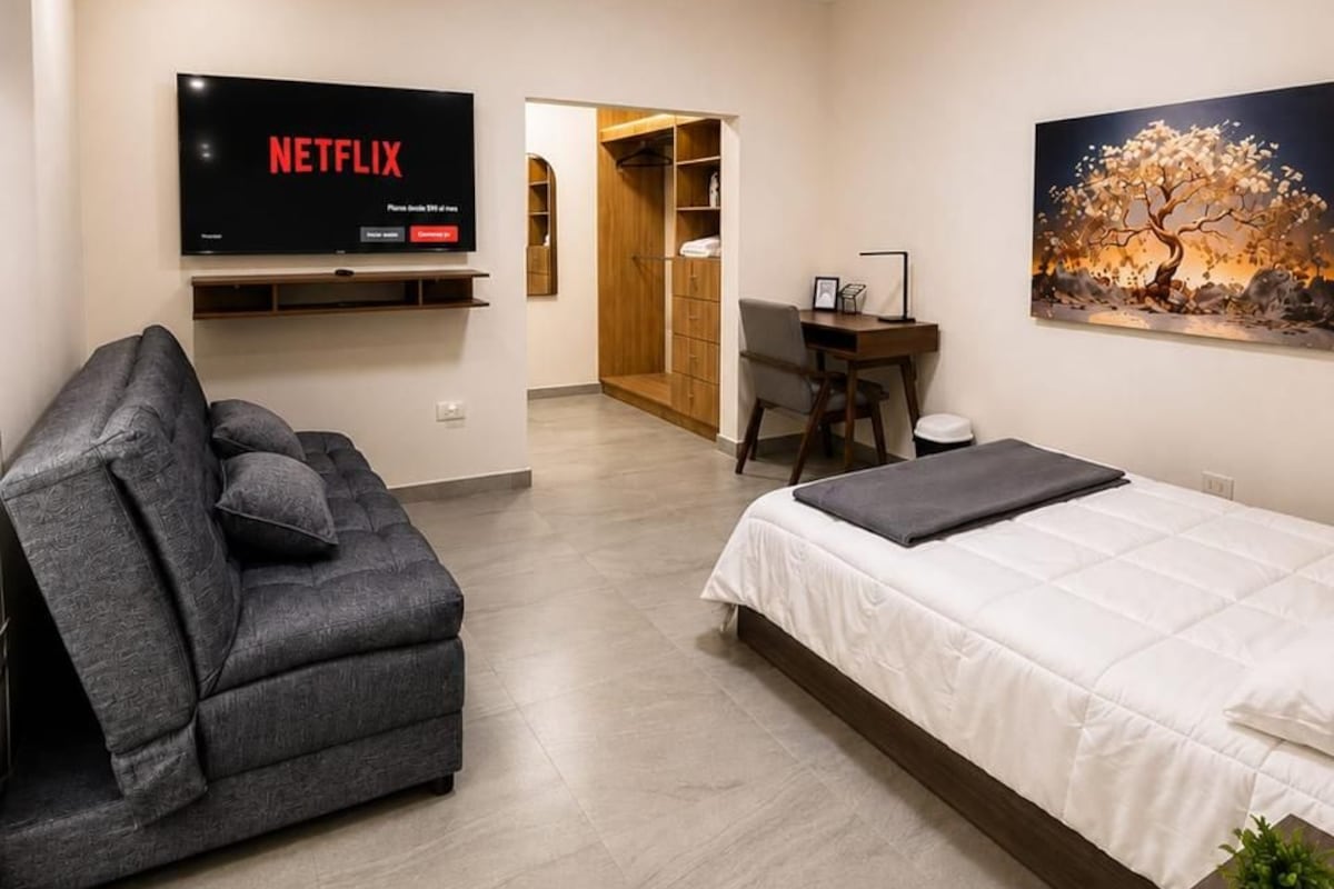 A modern hotel room with a bed, a sofa, a desk, and a TV displaying Netflix.