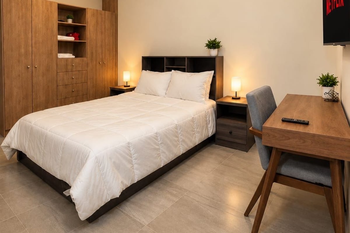 A bedroom with a bed, a desk, a chair, a wardrobe, a TV, and an air conditioning unit.