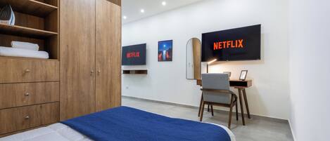 A modern hotel room with a bed, wooden wardrobe, desk, and two large TVs displaying the Netflix logo.
