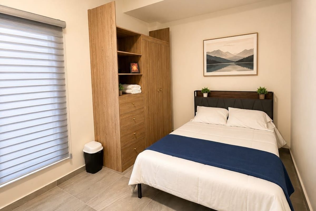 A bedroom with a bed, a wooden wardrobe, a picture on the wall, and a window with blinds.
