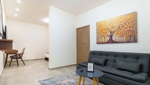 A modern living room with a grey sofa, a small round table with a QR code, a painting of a tree, and a wooden door. - Bella Terra (Hermosillo)