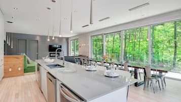 A modern kitchen with a large island, dining area, and large windows with a forest view.