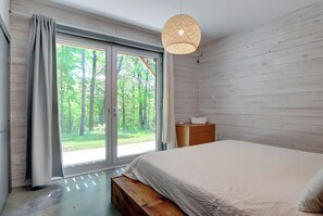 Une chambre à coucher comprenant un lit, un banc en bois, une table de chevet, un luminaire suspendu et une porte-fenêtre coulissante donnant sur un balcon avec vue sur les arbres.