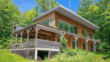 A modern wooden house with a large deck, surrounded by lush greenery.