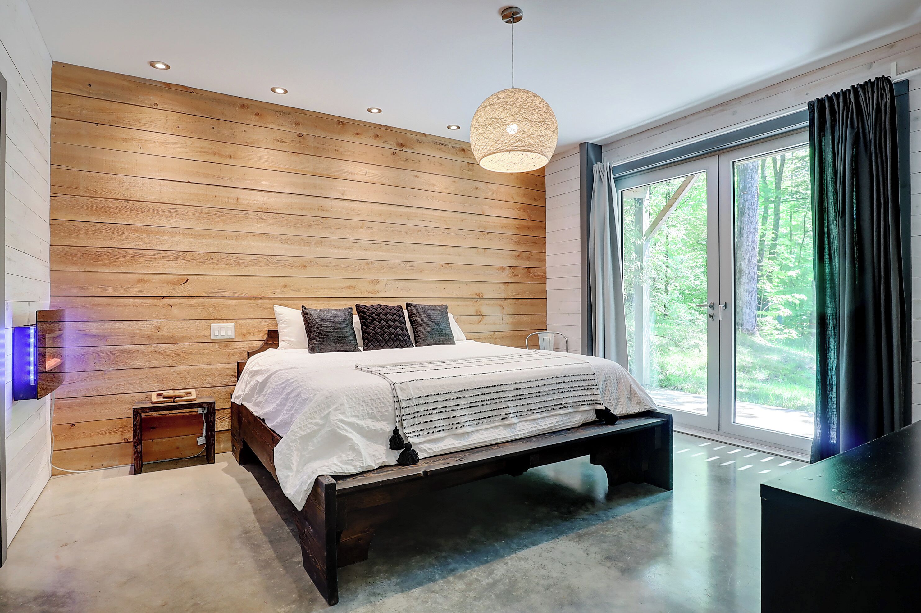 Een moderne slaapkamer met een houten wand, een bed met wit beddengoed, een hanglamp en een groot raam met gordijnen.