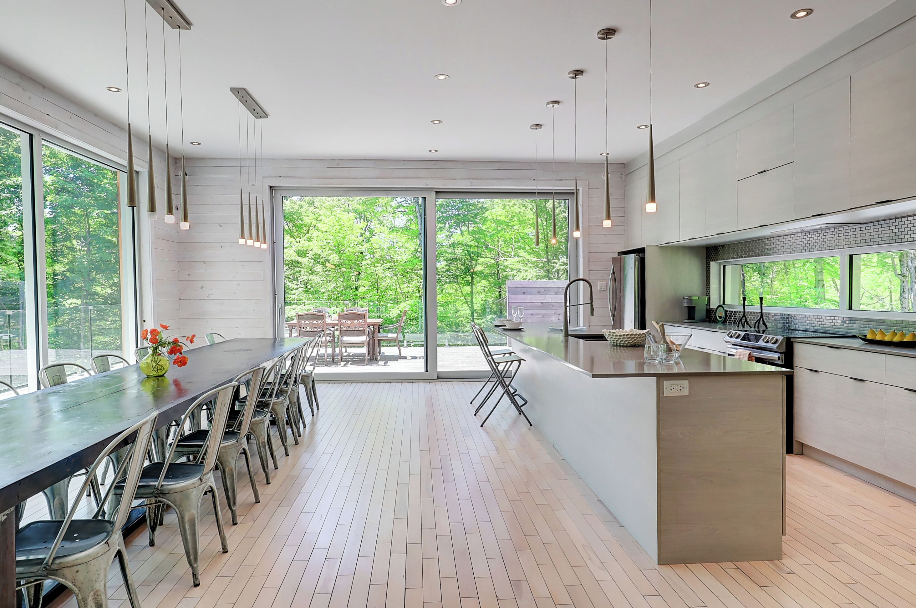 A modern kitchen with a large island, dining area with chairs, and large windows offering a view of greenery.