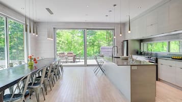 A modern kitchen with a large island, dining area with chairs, and large windows offering a view of greenery.