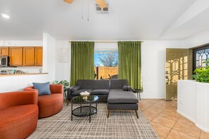 A living room with a sofa, armchair, and coffee table, a kitchen area with cabinets, and a view through green curtains. - Secluded Retreat: Private, Beautiful Home Wow (Tucson)