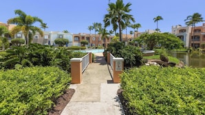 A pathway leading to a pool area surrounded by palm trees and a water body, with buildings in the background. - Fairway Courts Luxury Studio (FC872B) (Humacao)
