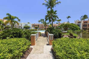 A pathway leading to a pool area surrounded by palm trees and a water body, with buildings in the background. - Fairway Courts Luxury Studio (FC872B) (Humacao)