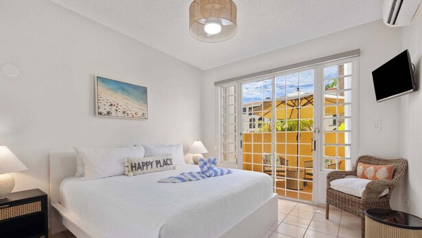 A bedroom with a bed, a television, a wicker chair, and a balcony with a view. - Fairway Courts Luxury Studio (FC872B) (Humacao)