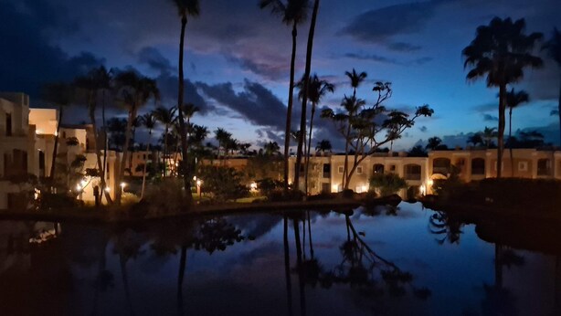 Vista nocturna de un complejo turístico con palmeras, un estanque reflectante y edificios iluminados.