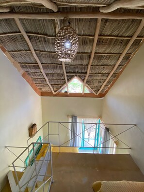 Ein Zimmer mit einem Strohdach, einer Hängelampe, einer Treppe mit Metallgeländer und einem Fenster mit Vorhängen.