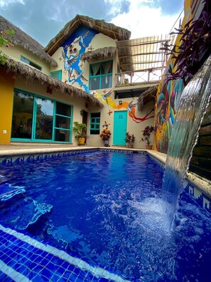 A swimming pool with a waterfall feature, surrounded by colorful murals and thatched-roof structures. - Casa Tropical Nibasan, beautiful garden and swimming pool (San Francisco)