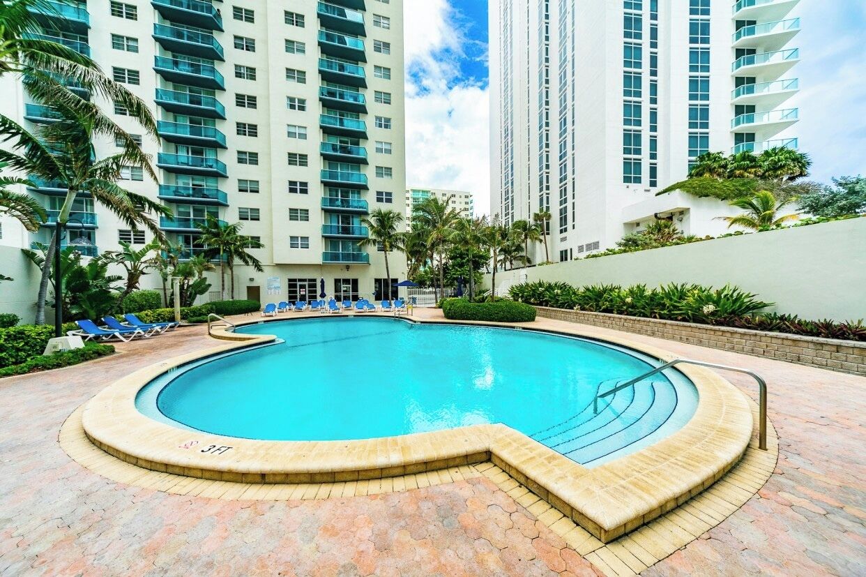 A swimming pool with a curved edge, surrounded by lounge chairs and palm trees, with high-rise buildings in the background.