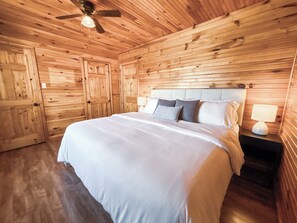Quarto com painéis de madeira, uma cama, um ventilador de teto e uma mesa de cabeceira com abajur.