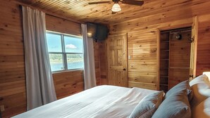 Een slaapkamer in een houten chalet met een bed, gordijnen, een plafondventilator en uitzicht op de natuur.