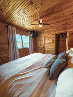 Un dormitorio tipo cabaña de madera con cama, cortinas, ventilador de techo y vista al exterior.