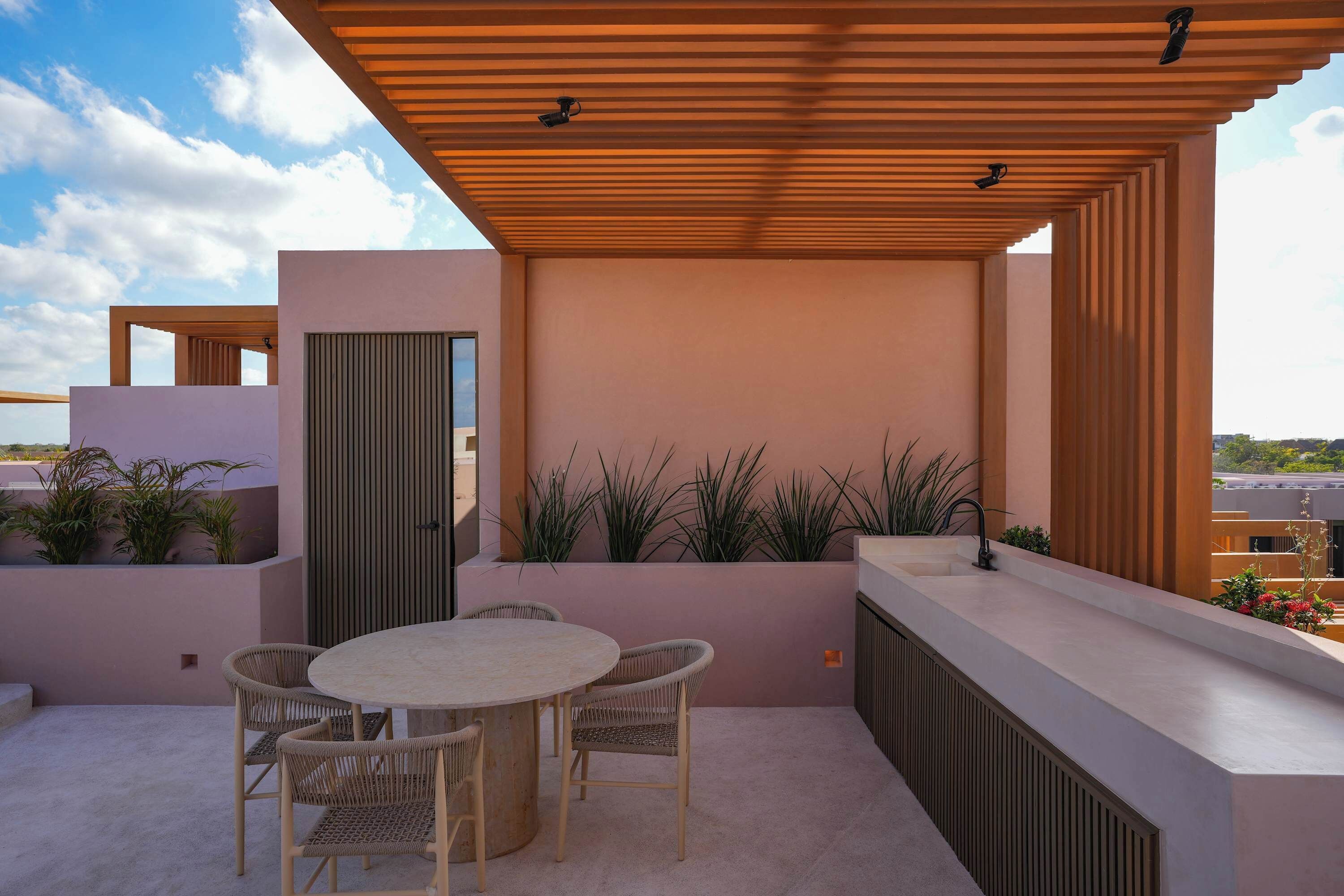 A rooftop terrace with a round table and chairs, a wooden pergola, and potted plants.