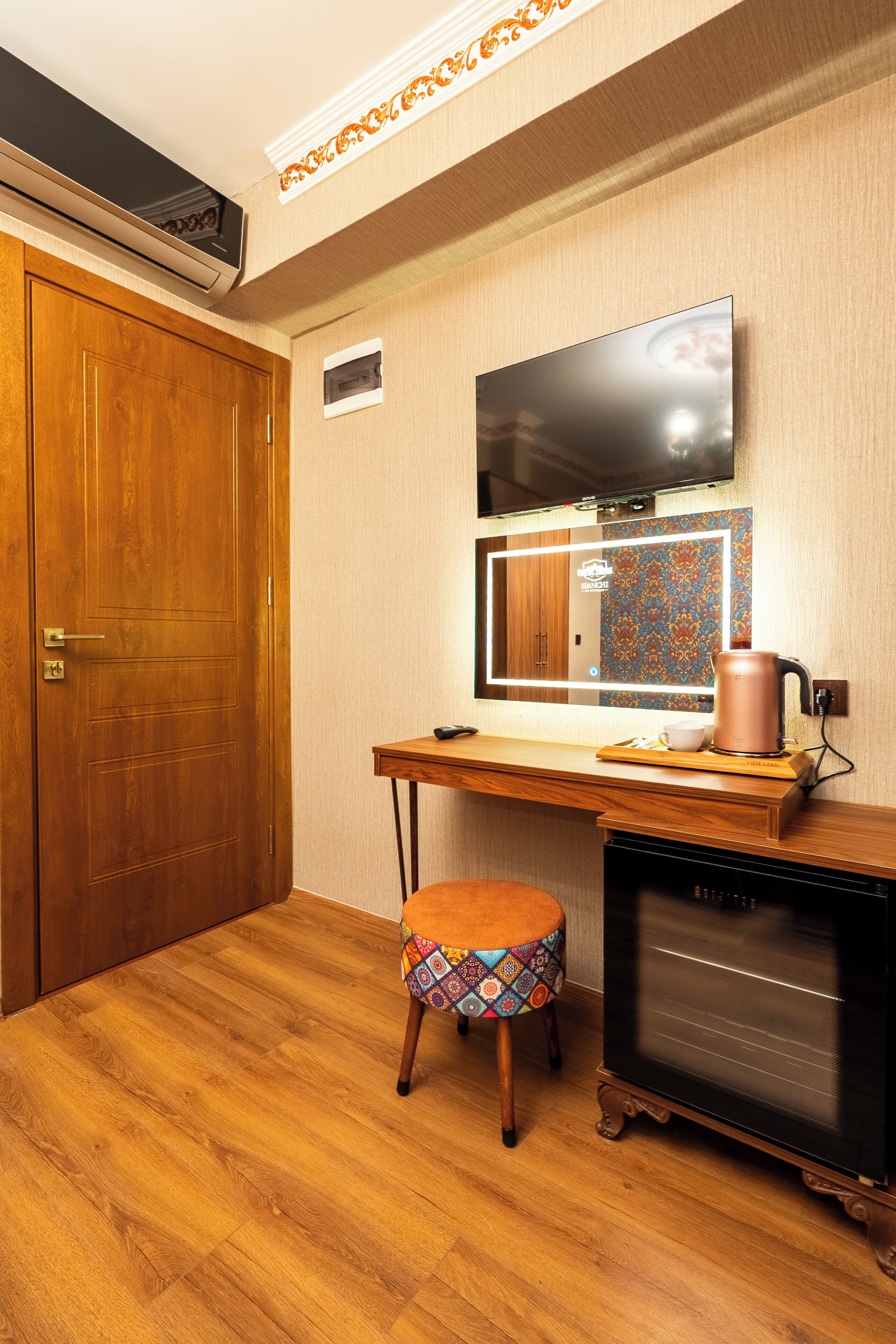 A room with a wooden door, a desk with a mirror, a television, a kettle, and a small stool.