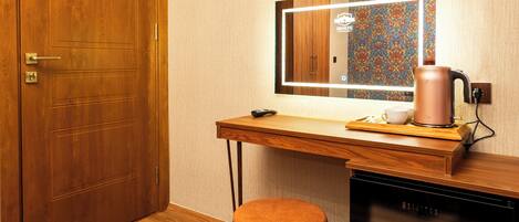 A room with a wooden door, a desk with a mirror, a television, a kettle, and a small stool.