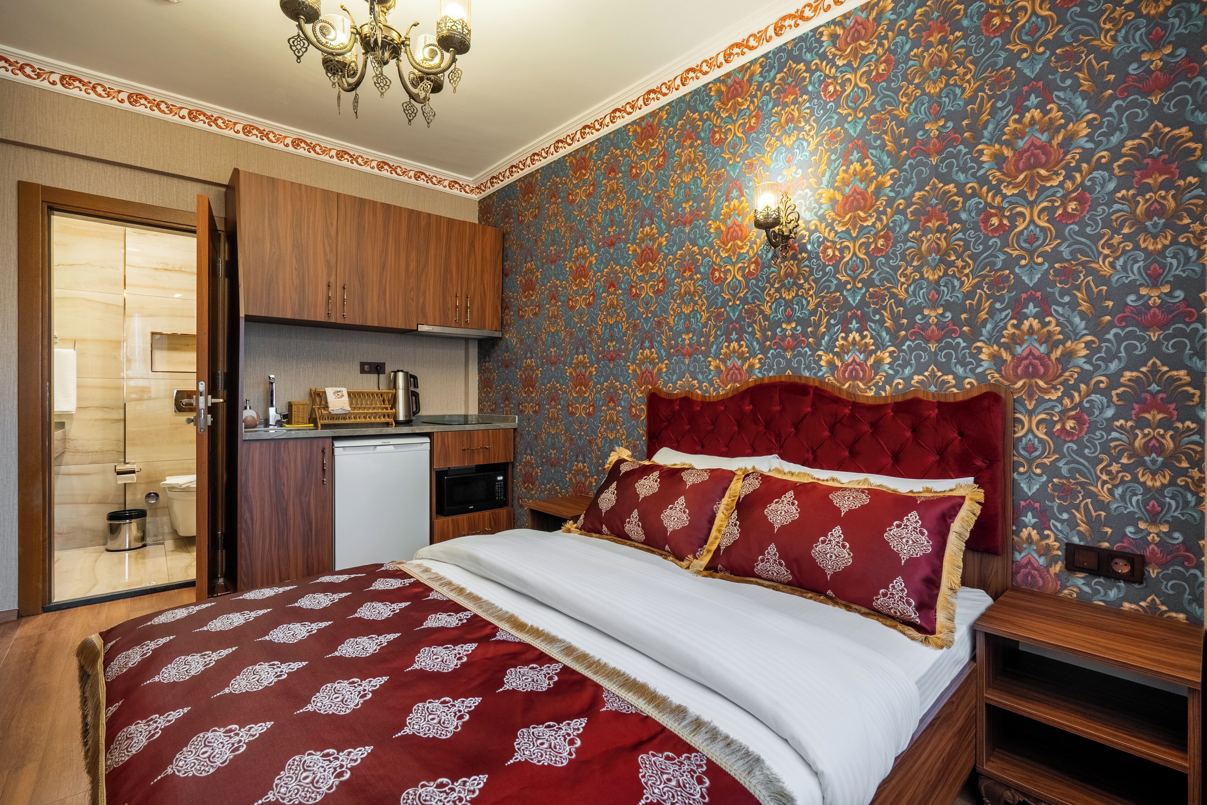 Un dormitorio con cama, cabecera tapizada en rojo, armario de madera, kitchenette pequeño y pared con estampado.