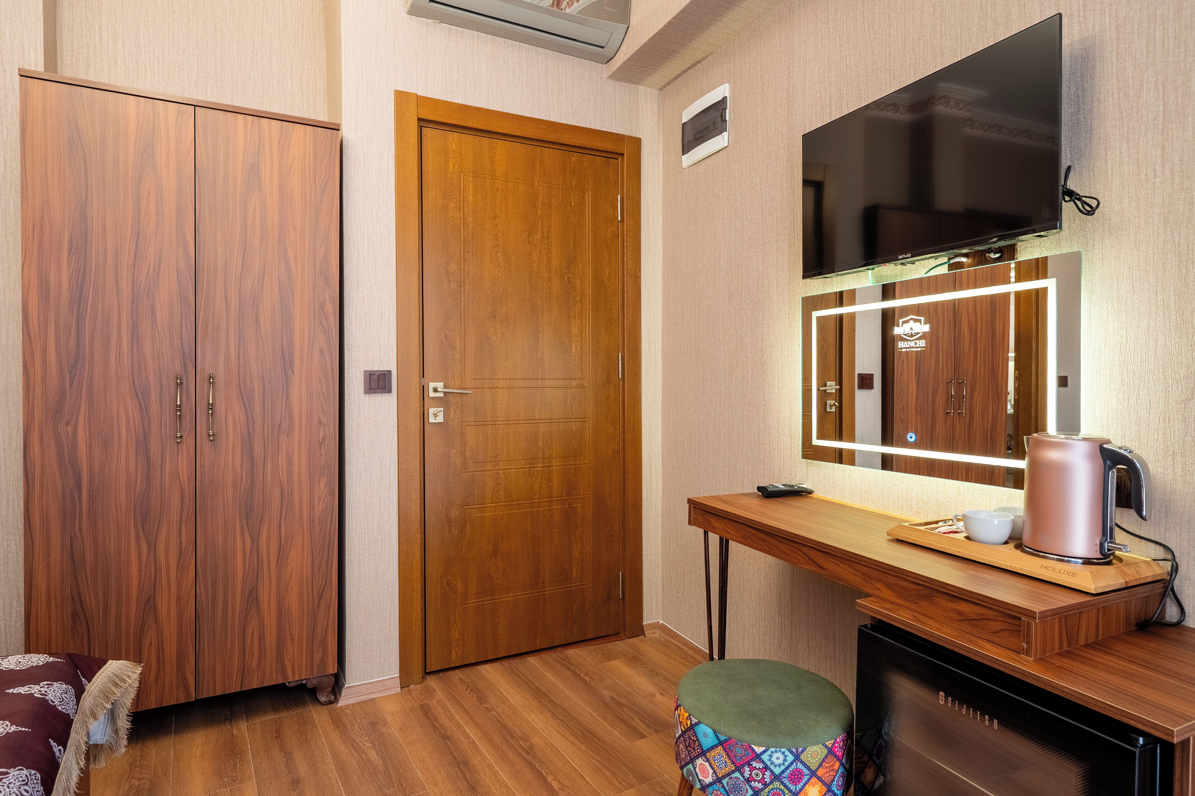 A room with a wooden wardrobe, a desk with a kettle, a television, and a sofa.