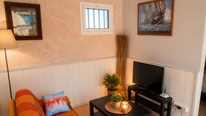 Ein Wohnzimmer mit einer Couch, einem Fernseher, einem kleinen Tisch mit einer Pflanze und einem Fenster.