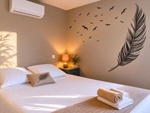Ein Schlafzimmer mit Bett, Kissen, einem Nachttisch mit Lampe, einer Wanddekoration mit Vögeln und einer Feder sowie einer Klimaanlage.