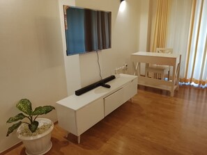 Ein modernes Wohnzimmer mit einem wandmontierten Flachbildfernseher, einem weißen TV-Schrank mit Fernbedienung, einem Esstisch mit Stühlen und einer Topfpflanze.