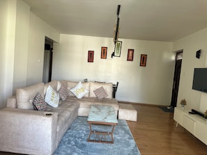 Ein Wohnzimmer mit einem beigen Sofa, gemusterten Kissen, einem Marmortisch, einem Deckenventilator, gerahmten Bildern an der Wand und einem Fernseher auf einem weißen Schrank.