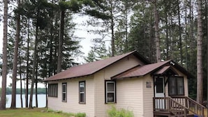 Eine kleine Holzhütte mit Veranda, umgeben von hohen Bäumen und einem Gewässer.