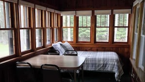 Ein Zimmer mit einem Bett, einem Tisch und Stühlen, große Fenster mit Blick auf Bäume.