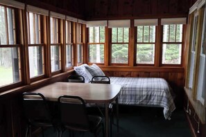 Ein Zimmer mit einem Bett, einem Tisch und Stühlen, große Fenster mit Blick auf Bäume.