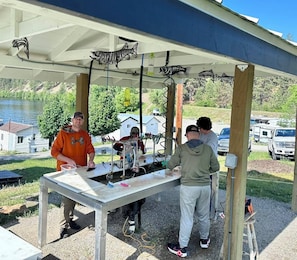 Un espacio al aire libre cubierto con una mesa, personas trabajando en un proyecto y un lago al fondo.