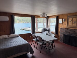 A room with a bed, a table with chairs, a fireplace, and a view of a lake with boats.