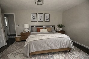 A bedroom with a bed, nightstands, a lamp, framed artwork, and a potted plant.