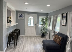 Una habitación con un sofá de color oscuro, una puerta blanca, una planta y un estante montado en la pared con un jarrón.