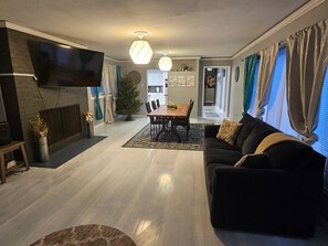 A living room with a flat-screen TV, a fireplace, a dining area with a table and chairs, and a sofa with cushions.