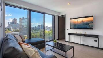 A modern living room with a large window, a black leather sofa, a coffee table, and a flat-screen TV mounted on the wall.