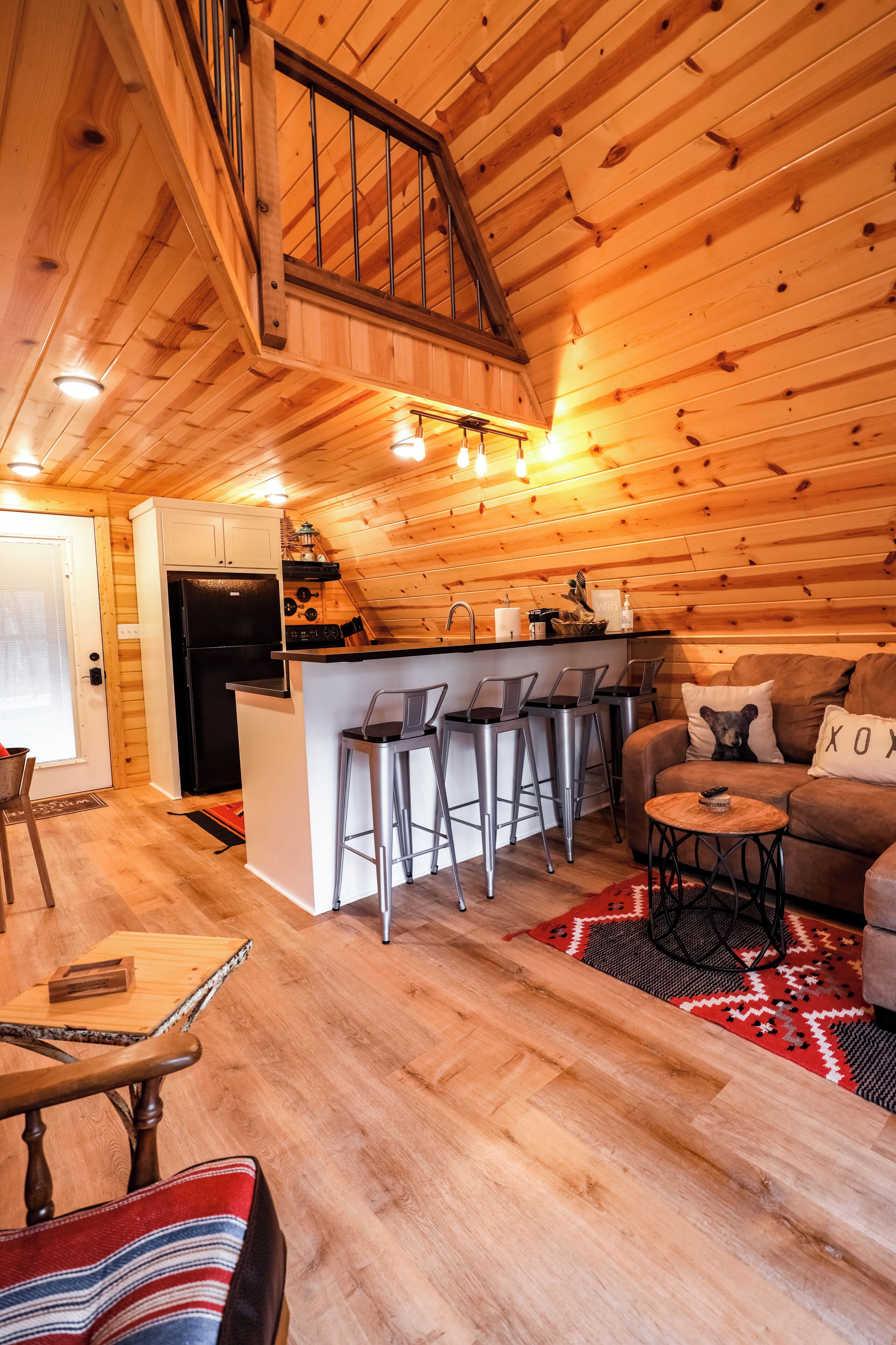 Un intérieur de chalet chaleureux avec un plafond en bois, un coin cuisine équipé d’un réfrigérateur, une table à manger avec des chaises et un coin salon avec un canapé et une table basse.