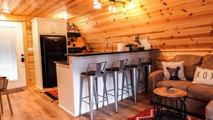 Un intérieur de chalet chaleureux avec un plafond en bois, un coin cuisine équipé d’un réfrigérateur, une table à manger avec des chaises et un coin salon avec un canapé et une table basse.