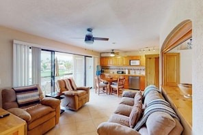 A living room with a sofa, chairs, a dining table, and a kitchen area with wooden cabinets and a ceiling fan. - Inviting Apartment Across From the Beach With Pool, Grill, Balcony & Golf Nearby (San José del Cabo)