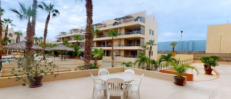 Terraza en la azotea con mesa y sillas, rodeada de plantas en macetas y palmeras, con vistas a un edificio con balcones.