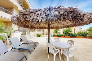 An outdoor seating area with a thatched umbrella, lounge chairs, and a round table. - Inviting Apartment Across From the Beach With Pool, Grill, Balcony & Golf Nearby (San José del Cabo)