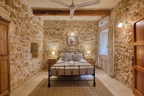 Une chambre avec un mur en pierre, un lit avec un cadre en métal, deux tables de chevet avec des lampes, un ventilateur de plafond et une porte en bois.