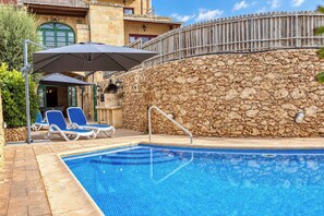 Une piscine aux carreaux bleus, un mur de pierre et deux chaises longues placées sous des parasols.
