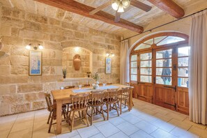 A stone-walled room with a wooden dining table and chairs, a large arched window, and a ceiling fan. - Tan Nannu 5BDR Farmhouse in Xaghra by PMI Malta (Ix-Xagħra)