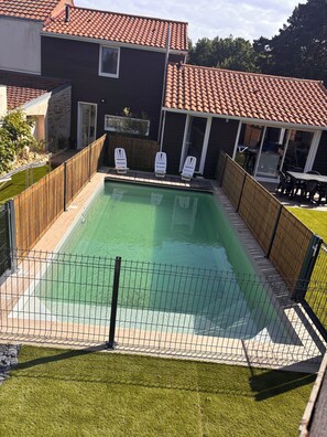 A rectangular swimming pool with a wooden deck and a fence, surrounded by a house with a tiled roof and a covered patio area. - Vrbo Property (Pornic)