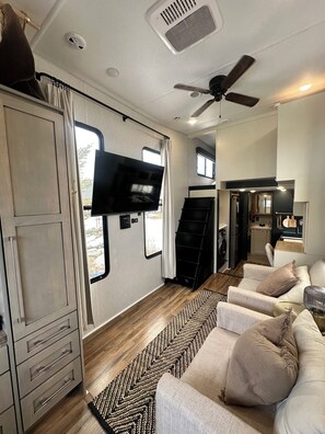 Interior compacto de uma RV, com televisão, ventoinha no teto, pequeno espaço de cozinha e sofá.